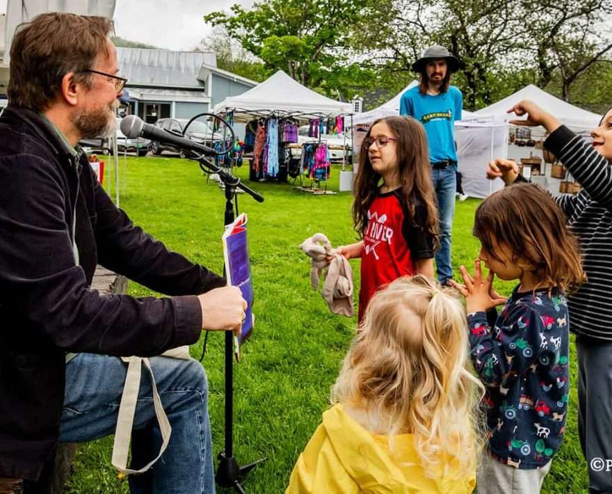 A man reading to a group of kids on the stage at a Vermont Farmers Market in Waitsfield